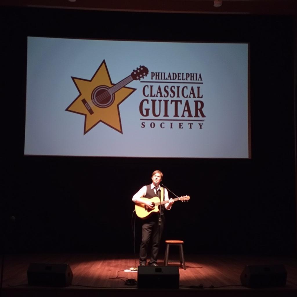 A packed auditorium watches a classical guitarist perform on stage with the PCGS logo in the background.